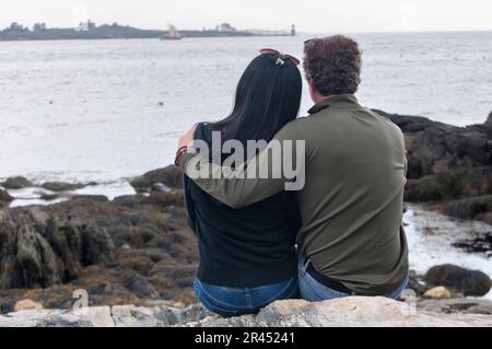 Ein Mann und eine Frau sitzen an einem bedeckten Tag am felsigen Ufer mit Blick auf die Ramininsel-Lichterstation. Stockfoto