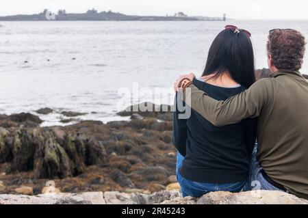 Ein Mann und eine Frau sitzen an einem bedeckten Tag am felsigen Ufer mit Blick auf die Ramininsel-Lichterstation. Stockfoto