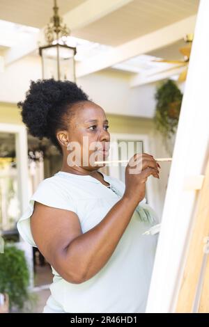 Fokussierte, glückliche afroamerikanische Seniorin, die zu Hause Leinwand auf Staffelei malt Stockfoto