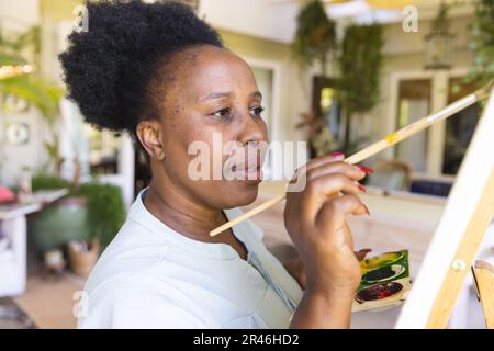 Fokussierte, glückliche, ältere afroamerikanische Frau, die zu Hause Leinwand mit Farbpaletten auf Staffelei hält Stockfoto
