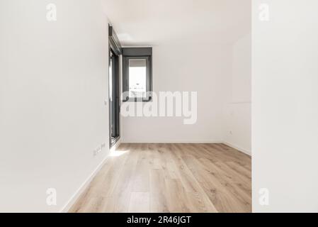 Ein leeres Schlafzimmer mit weißen Steckdosen an der Wand, einem Fenster und einer dunklen Aluminiumtür zu einer Terrasse und Holzfußboden und weiß bemalten Wänden Stockfoto