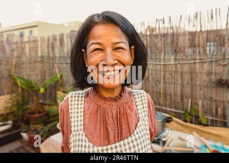 Eine glückliche Thai-Frau, die vor der Kamera lächelt, während sie das Rezept auf der Terrasse des Hauses zubereitet Stockfoto