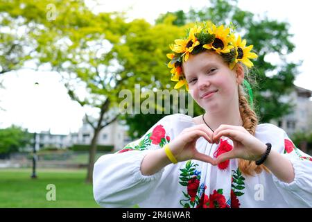 Eine ukrainische Frau in einem bestickten Hemd mit einem Sonnenblumenkranz auf dem Kopf macht ein Herz mit ihren Händen Anerkennung Anerkennung Freude Nadezhda angenehme Emotionen rothaarige Schönheit auf der Straße Stockfoto