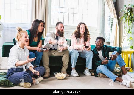 Mehrere ethnische Gruppen von Freunden, die Spaß beim Spielen von Videospielen haben. Lachen und Lächeln auf der Couch mit Steuerungen in der Hand Stockfoto