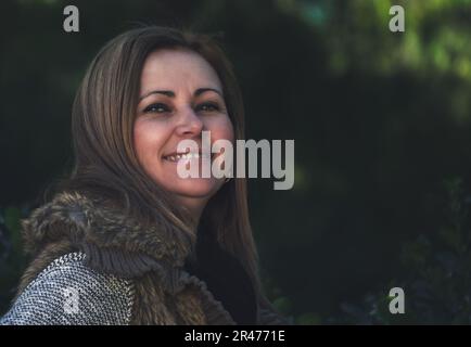 Porträt einer lächelnden blonden Frau mittleren Alters in der Natur, mit unscharfem dunkelgrünen Hintergrund. Lifestyle-Konzept. Stockfoto