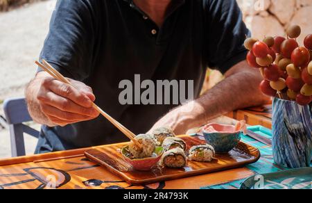 Ein Mann sitzt an einem Tisch und isst Sushi mit einem Paar Essstäbchen in der rechten Hand. Stockfoto