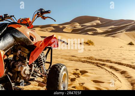 Quadbike-Fahrzeug in Orange und Schwarz parkt in der Sahara-Wüste, selektiver Fokus im Vordergrund, goldgelbe Sanddünen und blauer Himmel. Stockfoto