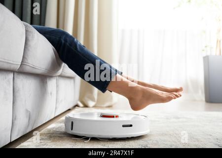 Der Roboter-Staubsauger führt die Reinigung im Wohnzimmer durch. Das Mädchen hob die Beine, um den Roboter-Staubsauger vorbeizulassen. Stockfoto