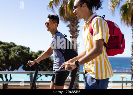 Ein glückliches, birassisches, schwules Paar, das Motorroller auf der Promenade am Meer fährt Stockfoto