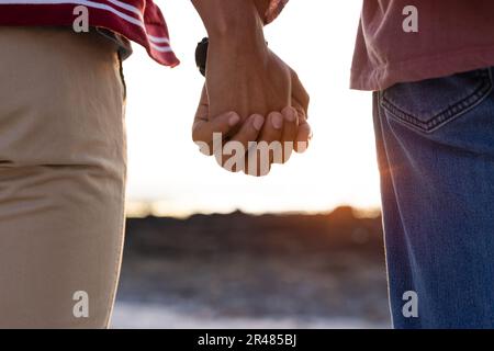 Mitten in einem birassischen, schwulen Paar, das bei Sonnenuntergang Hände am Strand hält Stockfoto