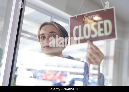 Fröhliche, weiße Friseurin, die das Schild an der Glastür des Friseursalons zu „Geschlossen“ wechselt Stockfoto