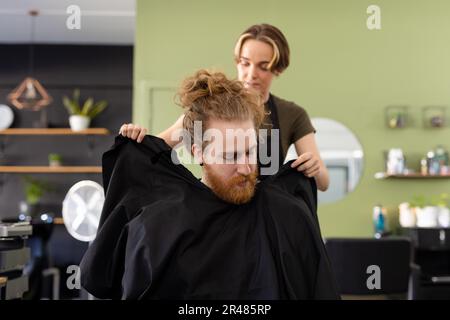 Eine weiße Friseurin, die männlichen Kunden im Friseursalon einen Umhang anzieht Stockfoto