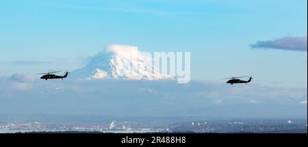 Zwei UH-60m Black Hawk Helikopter, die der Charlie Company zugewiesen wurden, 2-158 Angriffs-Helikopter-Bataillon, 16. Kampfluftfahrtbrigade fliegen am 8. Februar 2023 über Puget Sound in Washington. Mount Rainier ist im Hintergrund sichtbar. Stockfoto