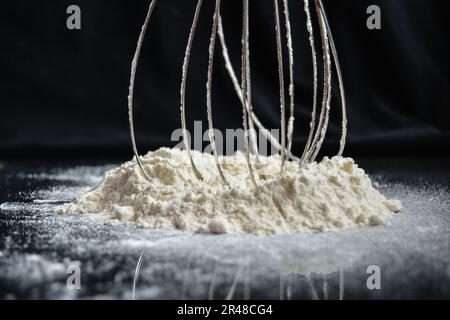 Ein Schneebesen aus rostfreiem Stahl, der in einem Hügel aus weißem Mehl vor einem festen Hintergrund eingetaucht ist Stockfoto