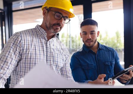 Verschiedene Geschäftsleute, die Architekturpläne haben, Tablets verwenden, über die Arbeit im Büro sprechen, unverändert Stockfoto