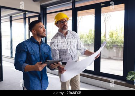 Verschiedene Geschäftsleute, die Architekturpläne haben, Tablets verwenden, über die Arbeit im Büro sprechen, unverändert Stockfoto