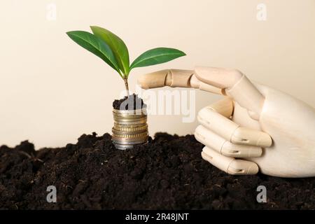 Münzstapel mit grüner Pflanze und hölzerner Schaufensterhand auf dem Boden vor beigefarbenem Hintergrund. Gewinnkonzept Stockfoto