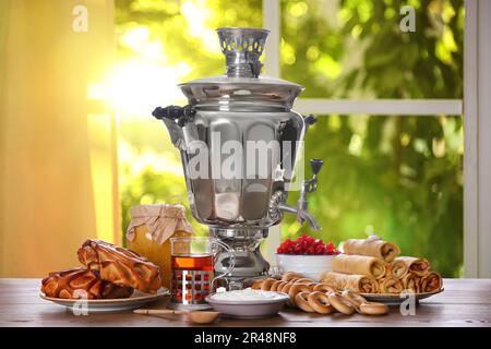 Traditioneller russischer Samovar und Leckereien auf einem Holztisch vor dem Fenster im Zimmer Stockfoto