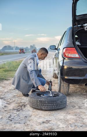 Ein Latino-Mann wechselt einen Platten Reifen am Straßenrand seines Wagens. Stockfoto