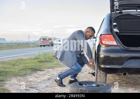 Ein Latino-Mann im Anzug wechselt einen Platten Reifen an seinem Auto am Straßenrand mit Kopierraum. Stockfoto