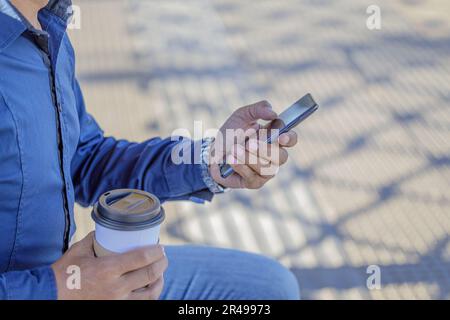 Detail der Hände eines Mannes mit einem Handy und einer Papptasse Kaffee. Stockfoto