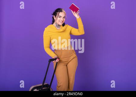 Junge glückliche Reisende mit Koffer, während sie ihren Pass vor lila Hintergrund vorzeigen. Speicherplatz kopieren. Studioaufnahme. Stockfoto
