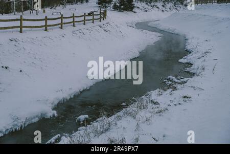 Ein ruhiger Bach schlängelt sich durch eine unberührte Winterlandschaft mit schneebedeckten Feldern Stockfoto