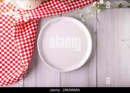 Kochhintergrund mit leerem weißem Teller und Picknick-Tischdecke auf rustikalem weißen Holztisch. Tabellenansicht mit Kopierbereich. Stockfoto