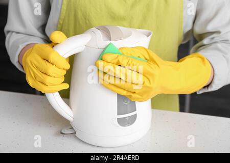 Frau in gelben Handschuhen, die elektrischen Wasserkocher auf einem weißen Tisch reinigt Stockfoto