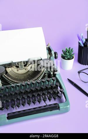 Vintage-Schreibmaschine mit Stifthalter, Brille und Hauspflanze auf lilafarbenem Hintergrund Stockfoto