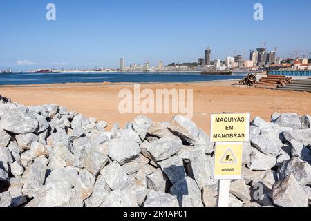 Blick auf die Küstenstadt Luanda, die neu aufgebaut wird, am Rand der Bucht. Baustellen, große Steinhaufen und Warnschild. Stockfoto