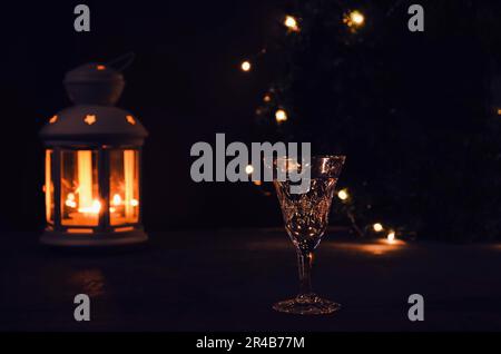 Glas mit Weißwein mit Girlande Lichter und Kerze Laterne auf dem Hintergrund. Dunkles Bild. Brennende Kerze. Goldenes Getränk im Glas. Wunderschön Stockfoto