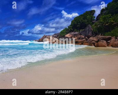 Eine ruhige Strandszene mit Wellen, die auf die felsige Küste schlagen. Grand Anse Beach, Seychellen. Stockfoto