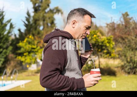 Ein erwachsener Mann hört Nachrichten auf seinem Handy und trinkt mit ihm, während er in seinem Garten spaziert. Home-Office-Konzept Stockfoto