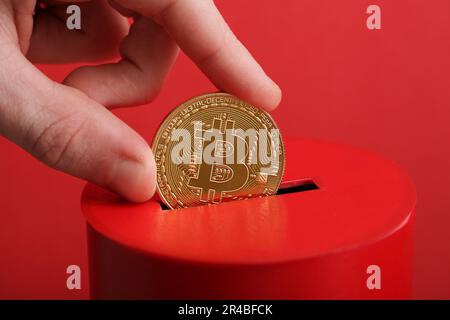 Männer stecken die goldene Münze mit Bitcoin-Symbol in den Schlitz der roten Spendenbox. Konzept Kryptowährung. Selektiver Fokus Stockfoto