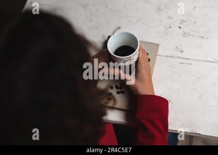 Eine lächelnde braune Frau, die einen roten Pullover und eine blaue Jeans trägt, genießt eine heiße Tasse Kaffee, während sie in einem gemütlichen Café sitzt Stockfoto