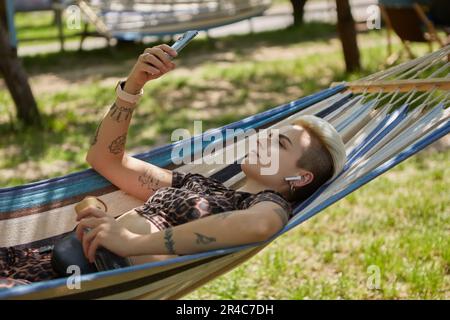 Stilvolle junge Frau mit kurzen gefärbten Haaren, die auf einer Hängematte in einem sonnigen grünen Park liegt, Musik hört und auf einem Smartphone im Internet surft. Vielfältig Stockfoto