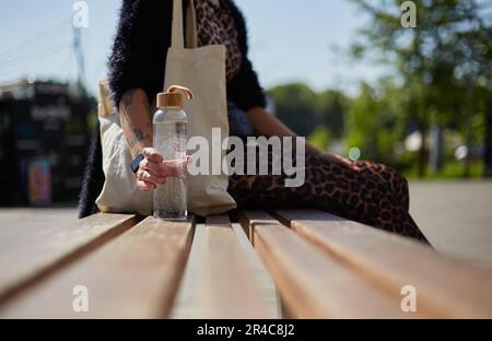 Junge Frau in Kleidung mit Leopardenmuster, die auf einer Bank sitzt, mit einer Glasflasche Wasser in der Hand und einer Baumwolltasche auf der Schulter. Nachhaltiger Lifestyle CO Stockfoto