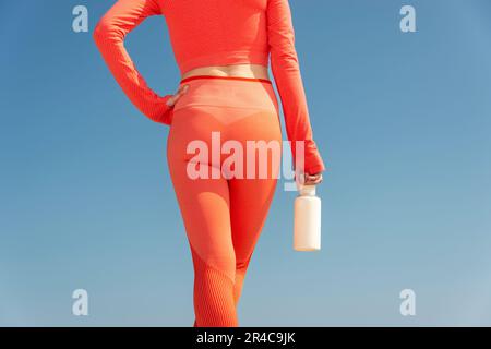 Rückansicht einer Sportlerin, die eine Flasche Wasser in der Hand hält und orangefarbene Aktivkleidung an einem blauen Himmel trägt. Stockfoto