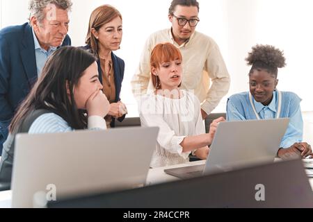 In einem modernen Büro sitzt eine rothaarige Frau und erklärt verschiedenen Kollegen, die an der Präsentation beteiligt waren, ein Projekt auf ihrem Laptop. Stockfoto