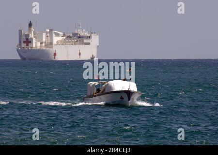 US Navy das militärische Sealift-Kommando-Krankenhausschiff USNS Mercy (T-AH 19) befindet sich am Horizont, während eines von Mercy's zwei Transportbooten sich dem Pier nähert, um Patienten, die nach der Lieferung eines Gesundheitswagens zurückkommen, abzusetzen. Stockfoto