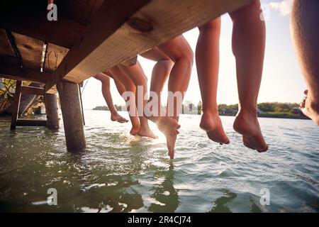 Junge Freunde sitzen am Dock und genießen gemeinsam im Urlaub baumelnde Beine im Wasser Stockfoto