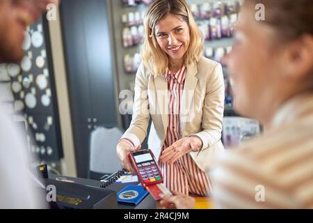 Wunderschöne junge Frau mit Kreditkartenleser und Interaktion mit einem jungen Ehepaar, das mit Kreditkarte bezahlt. Einkaufen, Lifestyle-Konzept. Stockfoto