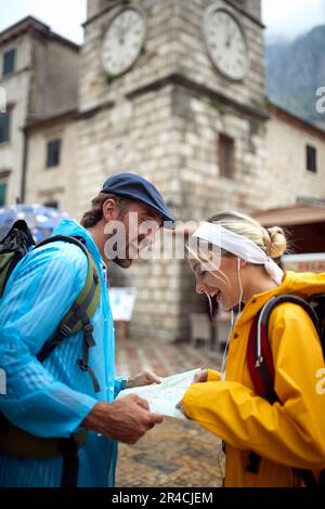 Glückliches Paar, das sich die Wegbeschreibung auf der Karte ansieht. Junger Mann und Frau in den Sommerferien in der Stadt, in Regenmänteln. Reisen, Zusammensein, Lifestyle-Konzept. Stockfoto