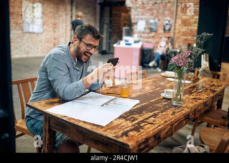 Fröhlicher, attraktiver junger Mann mit Brille und Kopfhörern im Café, der sein Smartphone lacht, Videoanrufe, Audioanrufe. Moderne Cafeteria im Hintergrund. Stockfoto