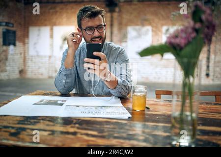 Junger attraktiver Mann sitzt im Café mit Kopfhörern über Smartphone. Moderner Café-Hintergrund. Stockfoto