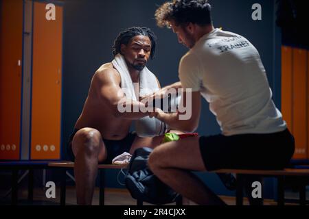 Ein persönlicher Trainer hilft dem Boxspieler, seine Boxhandschuhe nach dem Training auszuziehen und sich in der Garderobe auszuruhen. Sport, Lifestyle-Konzept. Stockfoto