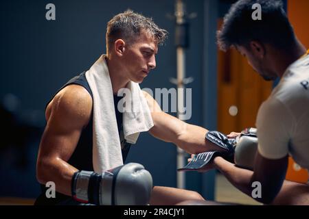 Junger muskulöser Boxspieler, der sich nach dem Spiel in der Garderobe ausruht, wobei der Trainer seine Handschuhe auszieht. Sport, Kampfsport, Lifestyle-Konzept. Stockfoto