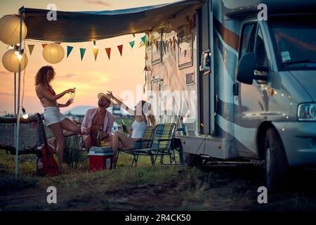 Drei Freunde, die draußen vor dem Wohnmobil anstoßen und Spaß haben. Sommeruntergang. Reisen, Urlaub, Zusammensein, Lifestyle-Konzept. Stockfoto