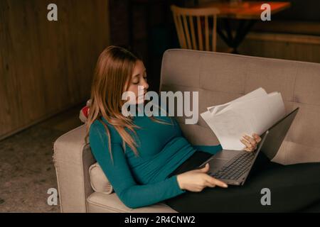 Wunderschöne blonde Frau, die Papierkram in der Hand hält, auf einem Sofa an der Kaffeebar liegt und auf dem Laptop arbeitet. Nahaufnahme Stockfoto